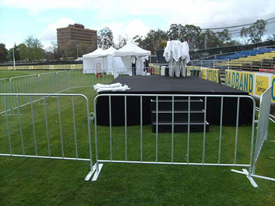 Crowd control barriers with flat feet are installed around a black performance stage and some tools.