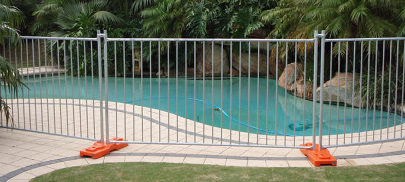 A galvanized temporary fence is installed at a pool. The temporary fence is supported by the orange plastic moulded feet.