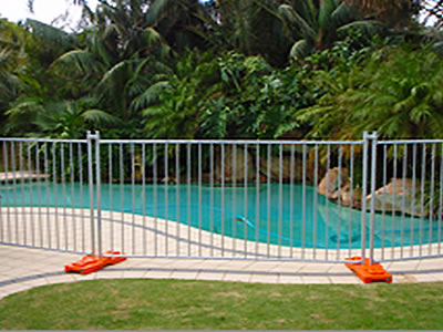 A galvanized temporary fence is installed at a pool. The temporary fence is supported by the orange plastic moulded feet.